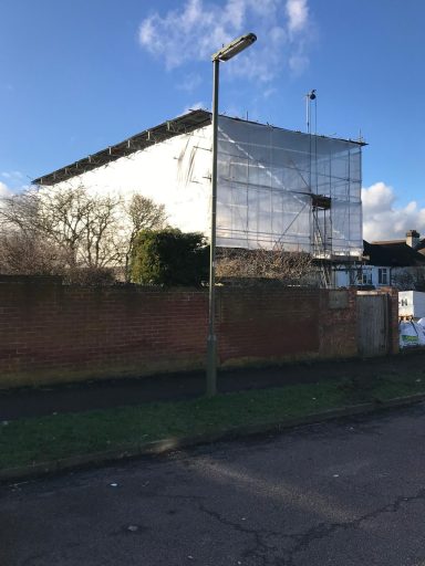 Modern building partially covered in scaffolding, surrounded by greenery and a lamppost.