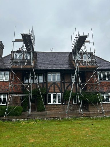 Scaffolding erected on a house with a dark roof and brick facade against a cloudy sky.